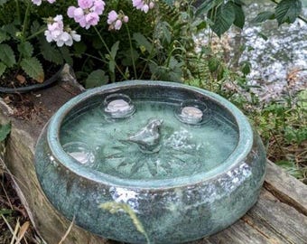 May include: A round, teal-colored birdbath with a metallic bird sculpture in the center. The birdbath holds water and several floating candles. The birdbath is placed on a weathered wooden surface, surrounded by greenery and flowers.
