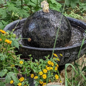 May include: A black, textured, spherical water fountain with water flowing from the top into a dark bowl. The fountain is surrounded by green plants and yellow flowers, creating a natural, outdoor aesthetic.