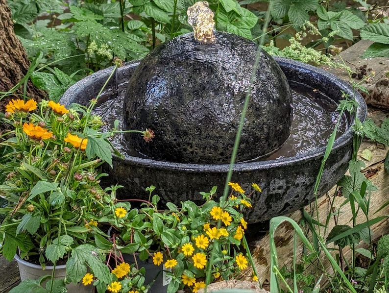 May include: A black, textured, spherical water fountain with water flowing from the top into a dark bowl. The fountain is surrounded by green plants and yellow flowers, creating a natural, outdoor aesthetic.