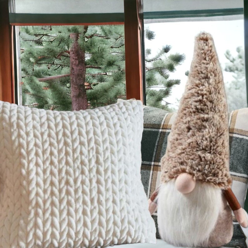 May include: A white knitted pillow with a cable knit pattern sits on a plaid couch next to a brown and white gnome with a long white beard.