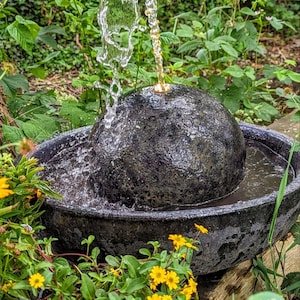 May include: A black stone water fountain with water cascading from a spherical top into a basin. The fountain is surrounded by green foliage and yellow flowers, creating a natural, outdoor aesthetic.