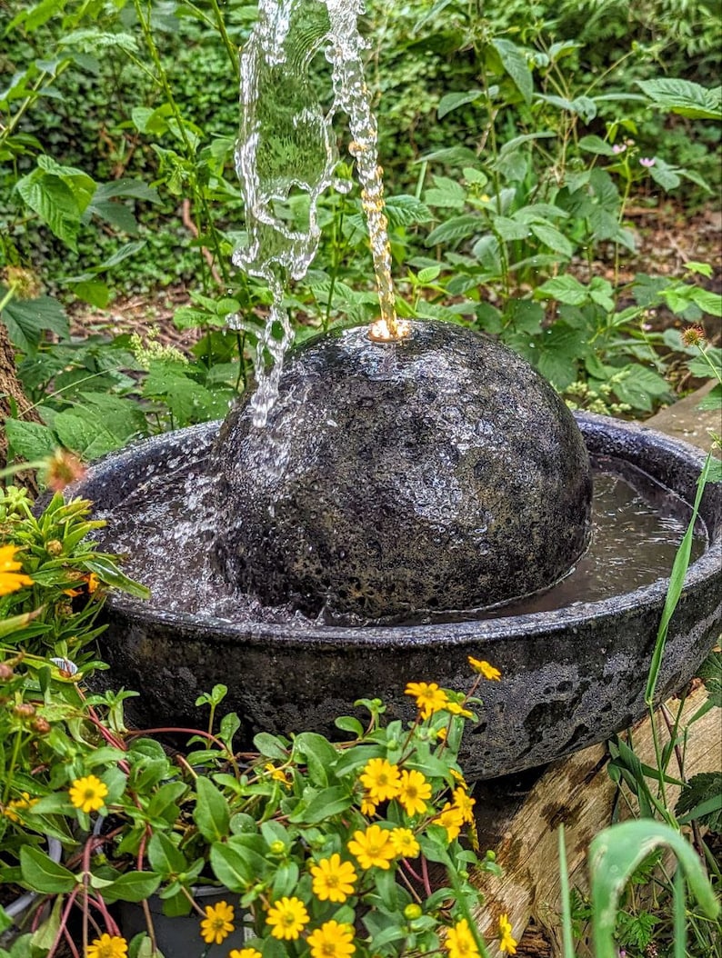 May include: A black stone water fountain with water cascading from a spherical top into a basin. The fountain is surrounded by green foliage and yellow flowers, creating a natural, outdoor aesthetic.