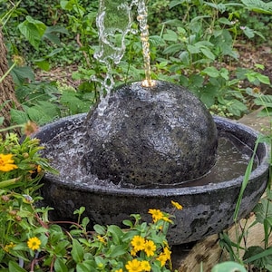 May include: A dark gray stone water fountain with water cascading from a spherical top into a basin. The fountain is surrounded by lush green foliage and yellow flowers, creating a natural, serene setting.