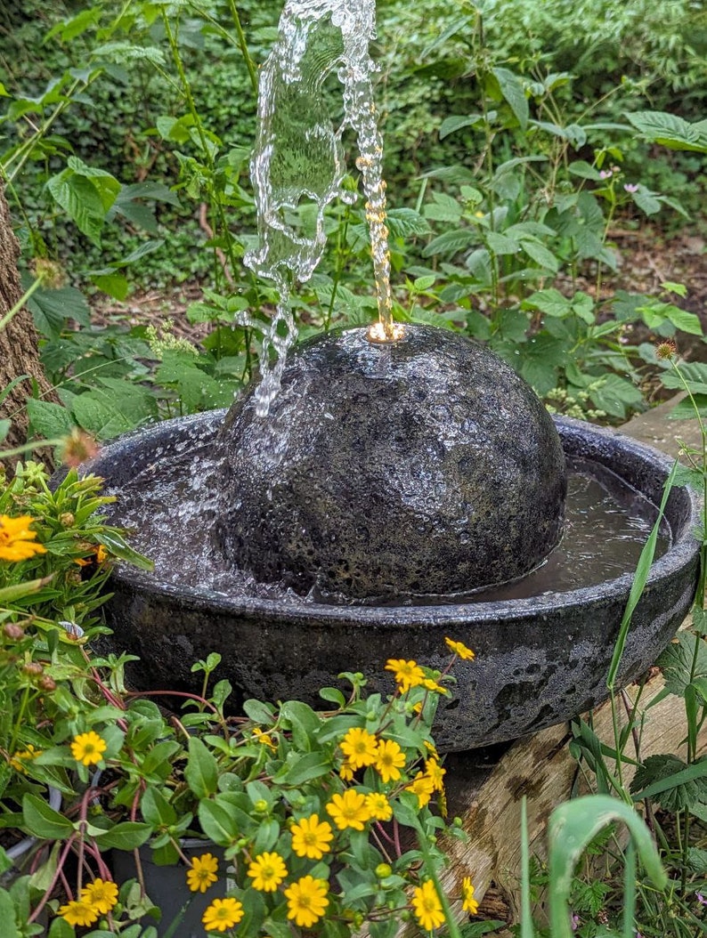 May include: A dark gray stone water fountain with water cascading from a spherical top into a basin. The fountain is surrounded by lush green foliage and yellow flowers, creating a natural, serene setting.