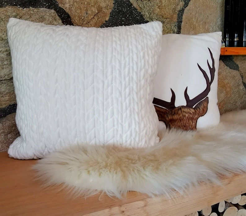 May include: Two white throw pillows, one with a cable knit texture and the other with a brown deer silhouette. The pillows are on a white faux fur rug.