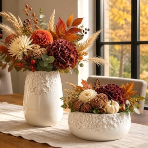 Op de afbeelding: Een hoge witte keramische vaas en een bijpassende lage witte keramische schaal, beide met een bloemrijk reliëfpatroon. De vaas bevat een arrangement van oranje dahlia's, witte chrysanten, rode bessen en gedroogde grassen. De schaal bevat donkerrode hortensia's, witte chrysanten en een kleine witte pompoen.