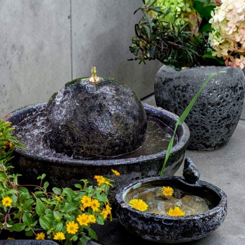 May include: A dark gray stone water fountain with a spherical top and a small, round basin. Water flows from the top into the basin. The fountain is surrounded by other stone bowls and a vase with flowers.