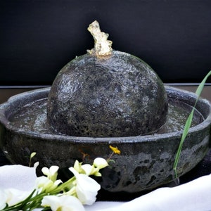 May include: A dark gray stone water fountain with a spherical top and a surrounding basin. Water flows from the top, creating a small cascade. White flowers and greenery are in the foreground, with a black background.