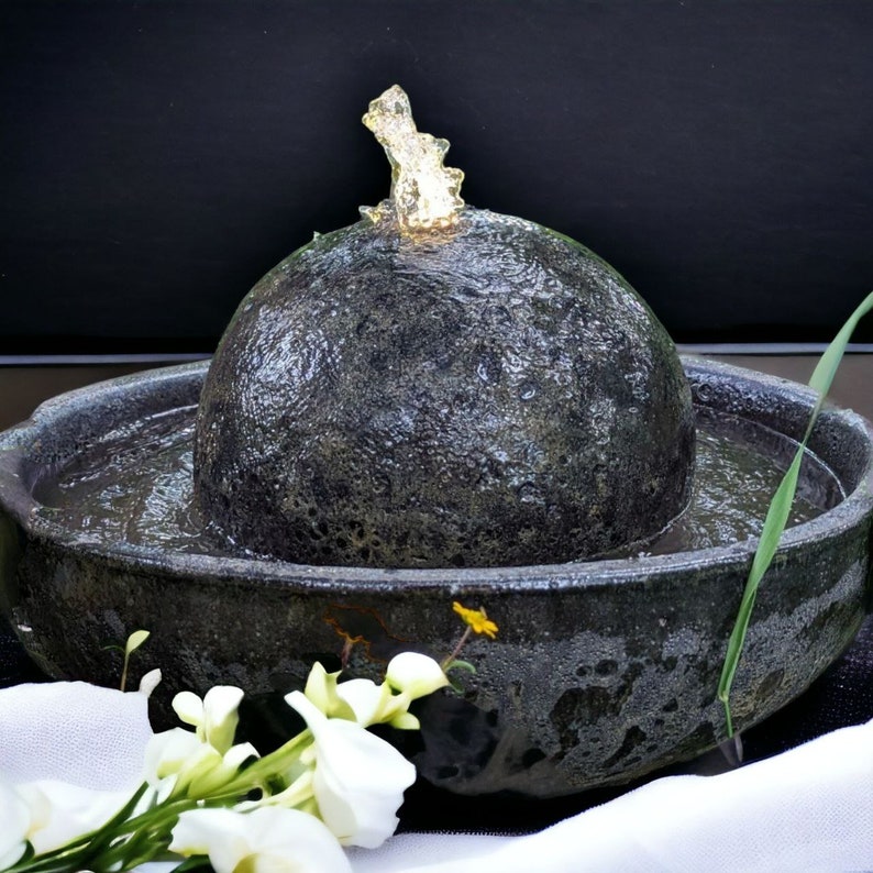 May include: A dark gray stone water fountain with a spherical top and a surrounding basin. Water flows from the top, creating a small cascade. White flowers and greenery are in the foreground, with a black background.
