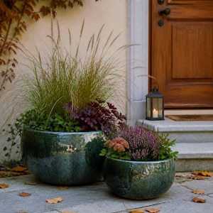 Può includere: Due vasi rotondi in ceramica color verde acqua lucido poggiano su un patio in pietra. Il vaso più grande contiene erbe ornamentali e fogliame viola, mentre quello più piccolo ospita erica viola e piccoli fiori rosa.