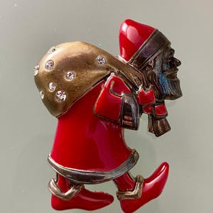 Puede incluir: Un broche de metal con forma de figura que viste un abrigo, gorro y botas de color rojo brillante con ribetes plateados. La figura lleva un saco grande y texturizado de color bronce sobre el hombro, decorado con varios pequeños brillantes transparentes.