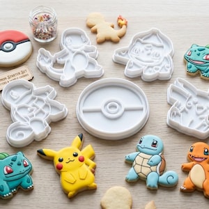 May include: A set of white plastic cookie cutters shaped like various characters and a circular ball, accompanied by decorated sugar cookies, a small jar of sprinkles, and a tag labelled Premium Pokemon Starter Set Handcrafted Cutters & Sample Cookies.