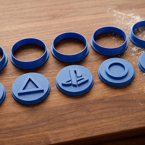 May include: A set of blue plastic cookie cutters featuring geometric shapes including a square, triangle, PlayStation logo, circle, and cross. The set includes five circular frames and five corresponding shape inserts, with one baked cookie shaped like a cross.