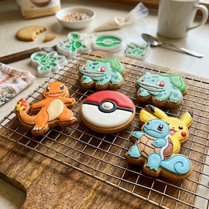 May include: A selection of decorated sugar cookies depicting Pokémon characters. The cookies include Charmander, Bulbasaur, Squirtle, and Pikachu, plus a Poké Ball design. The cookies are arranged on a wire rack over a wooden chopping board.