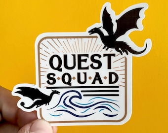May include: White sticker with a brown border and a sunburst design. The sticker says "QUEST SQUAD" in black text. There are two black silhouettes of dragons and a blue wave design.