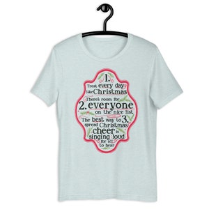Op de afbeelding: Lichtblauw T-shirt met een rood en groen grafisch ontwerp. De grafiek bevat een feestelijke boodschap over het behandelen van elke dag als Kerstmis. De tekst luidt: "Behandel elke dag als Kerstmis. Er is ruimte voor iedereen op de nette lijst. De beste manier om kerstvreugde te verspreiden is hardop te zingen zodat iedereen het kan horen."