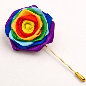 Op de afbeelding: Een reversspeld met een stoffen roos gemaakt van gelaagd lint in een regenboogkleurvolgorde van paars, blauw, lichtblauw, groen, geel, oranje en rood, bevestigd aan een lange, dunne, goudkleurige metalen speld met een bijpassende dop.