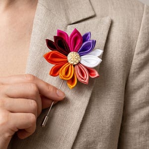 May include: A multi-colored fabric flower lapel pin featuring petals in shades of deep red, orange, yellow, pink, purple, and white. The center of the flower is a circular gold-toned metal button with an embossed pattern on a long pin.