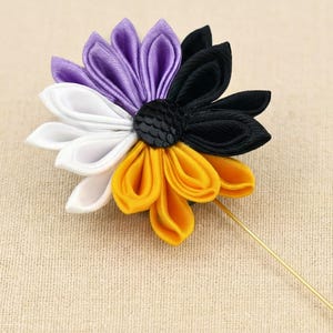 Könnte beinhalten: Eine Anstecknadel mit einer Stoffblume, deren Blütenblätter in vier verschiedenen Farbabschnitten angeordnet sind: Violett, Schwarz, Weiß und Gelb. Die Mitte der Blume ist eine kleine, runde, schwarze, strukturierte Scheibe. Eine dünne, gerade goldfarbene Metallnadel ragt heraus.