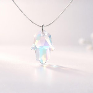 May include: A clear, iridescent crystal Hamsa hand pendant suspended from a thin silver-toned chain. The pendant features a faceted, multi-sided surface that reflects light, creating a spectrum of soft rainbow colours across its translucent body.