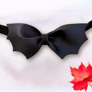 May include: A black bow tie featuring wing-shaped edges, attached to a thin black strap. The bow tie is constructed from a smooth, matte material and is positioned horizontally across the frame, with a small cluster of bright red maple leaves below.