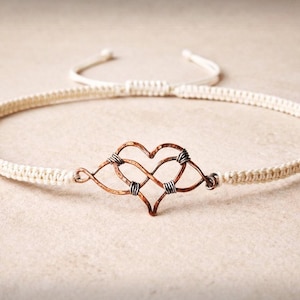 Könnte beinhalten: Cremefarbenes Makramee-Armband mit einem kupferfarbenen herzförmigen Anhänger mit Unendlichkeitszeichen. Das Armband hat einen verstellbaren Schiebeknotenverschluss. Schmuck.