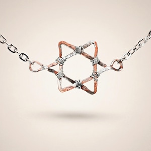 Puede incluir: Un collar hecho a mano con un colgante en forma de estrella. El colgante está hecho de alambre de cobre y plata, con una cadena de plata. La estrella tiene un diseño geométrico, con el alambre de cobre mostrando un tono marrón rojizo. El collar está suspendido sobre un fondo neutro.