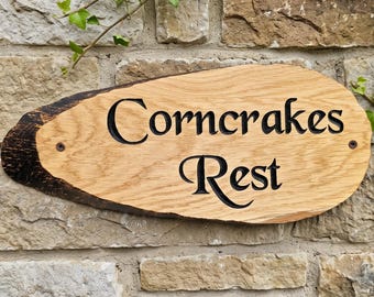 May include: A live edge wooden sign with a natural bark border on the left side. The light brown wood features the text "Corncrakes Rest" engraved in a dark, serif font. Two small metal screws are visible on the left and right sides of the sign.
