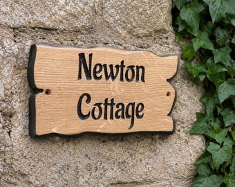 Op de afbeelding: Een rechthoekig houten bord met een natuurlijke lichtbruine houtnerfafwerking en donkerbruine randen. De tekst Newton Cottage is in het midden gegraveerd in een zwart schreeflettertype. Twee kleine ronde gaten zijn aan de zijkanten zichtbaar.