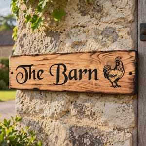 May include: A rectangular wooden sign with a natural light brown wood grain and charred edges. The sign features the black text "The Barn" in a decorative font on the left side and a black line art illustration of a rooster on the right side.