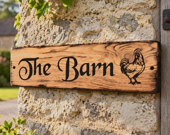 May include: A rectangular wooden sign with a natural light brown wood grain and charred edges. The sign features the black text "The Barn" in a decorative font on the left side and a black line art illustration of a rooster on the right side.