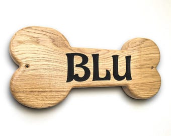 Op de afbeelding: Houten hondenbotvormig bord met de naam "BLU" in zwarte letters. Het bord is gemaakt van lichtgekleurd hout met zichtbare nerf en heeft een gladde afwerking. De botvorm heeft afgeronde uiteinden.