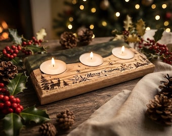 May include: A rectangular wooden candle holder with a dark resin accent, featuring three lit tea light candles. The wood is engraved with the text Merry Christmas, a deer, and pine trees. The piece is surrounded by holly berries and pine cones.