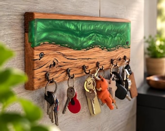 May include: A rectangular wall-mounted key holder made from a piece of natural wood with a prominent grain and a thick, translucent emerald green resin inlay along the top edge. It features six black metal hooks holding various metal key rings.