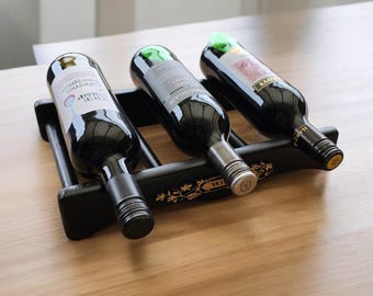 May include: Three wine bottles are displayed in a black wooden wine rack. The bottles are all different brands and have labels with gold lettering. The rack has a decorative design with gold accents.