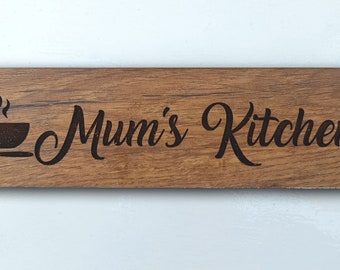 Op de afbeelding: Een houten bord met de tekst "Mum's Kitchen" in een cursief schrift. Links van de tekst staat een kleine zwart-witte illustratie van een stomende mok en een lepel.
