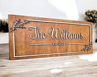 Op de afbeelding: Een houten bord met een wit frame en de tekst "The Williams Est. 2020" in een cursief schrift. Het bord is versierd met twee kleine, bladerrijke takken aan elke kant.