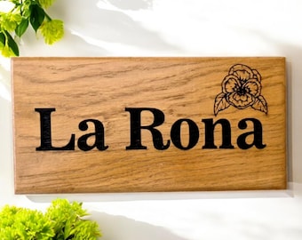 Op de afbeelding: Een houten bord met de naam "La Rona" gegraveerd in zwarte letters. Rechtsboven op het bord staat een klein zwart-wit bloemenmotief.