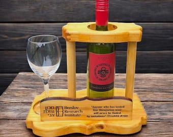 Op de afbeelding: Een houten houder voor een wijnfles en een glas met een citaat over Hongaarse wijn. De houder heeft een klein bordje met de tekst "Breslov Research Institute" en de Hebreeuwse tekst "מכון נחלת צבי".