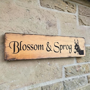 Puede incluir: Un letrero rectangular de madera con las palabras "Blossom & Sprog" en escritura negra y una silueta de burro negro. El letrero est&aacute; montado en una pared de piedra. La madera tiene vetas naturales y un borde oscuro.