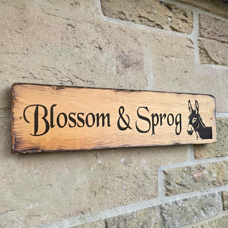 Puede incluir: Un letrero rectangular de madera con las palabras "Blossom & Sprog" en escritura negra y una silueta de burro negro. El letrero est&aacute; montado en una pared de piedra. La madera tiene vetas naturales y un borde oscuro.