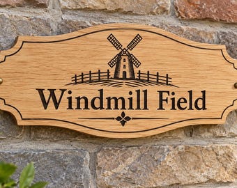 May include: A light brown wooden sign with a decorative scalloped edge, featuring a black engraved graphic of a windmill in a field above the text Windmill Field and a small floral icon, secured with two brass-colored metal screws on each side.