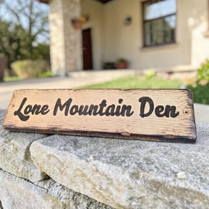 Puede incluir: Un letrero de madera rectangular con el texto grabado "Lone Mountain Den". El letrero tiene una superficie marr&oacute;n claro con bordes m&aacute;s oscuros y est&aacute; montado en un muro de piedra. Es probable que el letrero sea para una casa o negocio.