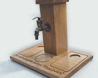Op de afbeelding: Houten tapdispenser met een bronzen kraan. De dispenser heeft een rechthoekige basis met uitgesneden cirkelvormige en rechthoekige inkepingen. De houten structuur heeft een verticale kolom met een kraan en een bovenstuk.