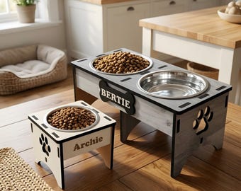 May include: Two elevated pet feeding stations made of wood. The larger gray station holds two stainless steel bowls and features a bone-shaped plaque with the name BERTIE. The smaller white station holds one bowl and features the name ARCHIE.