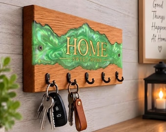 May include: A rectangular wooden key holder featuring a central band of swirling green resin. The text HOME SWEET HOME is printed in gold lettering across the resin. Five black metal hooks are attached to the bottom, with two sets of keys hanging.