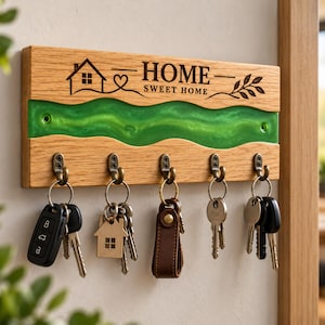 Può includere: Un portachiavi rettangolare in legno con un inserto orizzontale ondulato in resina verde brillante. Il legno è inciso con l'icona di una casa, un cuore, un rametto di foglie e il testo "HOME SWEET HOME". Cinque ganci in metallo bronzo reggono chiavi.