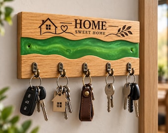 May include: A rectangular wooden key holder featuring a horizontal wavy inlay of bright green resin. The wood is engraved with a house icon, a heart, a leaf sprig, and the text "HOME SWEET HOME". Five bronze-toned metal hooks hold various keys.