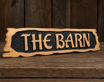 Op de afbeelding: Een rechthoekig houten bord met een natuurlijke rand, voorzien van de tekst THE BARN in reliëf uitgesneden. De letters zijn lichtbruin en steken af tegen een donkere, getextureerde, verzonken achtergrond die de houtnerf benadrukt.