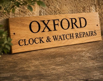 Op de afbeelding: Een rechthoekig lichtbruin houten bord met donkere gegraveerde letters met de tekst OXFORD CLOCK & WATCH REPAIRS. Het bord heeft een natuurlijke houtnerfstructuur en bevat twee kleine voorgeboorde gaten aan de zijkanten voor bevestiging.